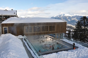 A modern building with a glass pool surrounded by snow, set against a mountainous backdrop.