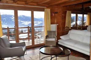 A cozy room with a white sofa, a grey armchair, and a round table. Large windows offer a view of snow-capped mountains.