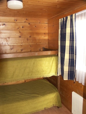 A cabin with wooden walls, a bunk bed with green bedding, a white radiator, and a window with striped curtains.