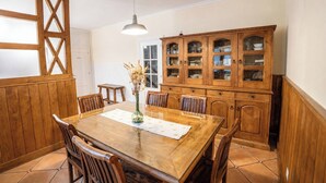 Een eetgedeelte met een houten tafel, stoelen, een vaas met gedroogde bloemen en een houten kast met glazen deuren.