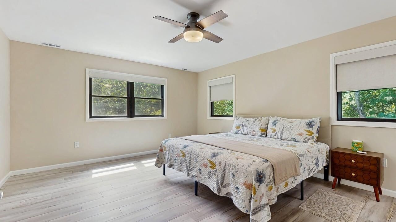 A bedroom with a bed, a nightstand, a ceiling fan, and two windows.