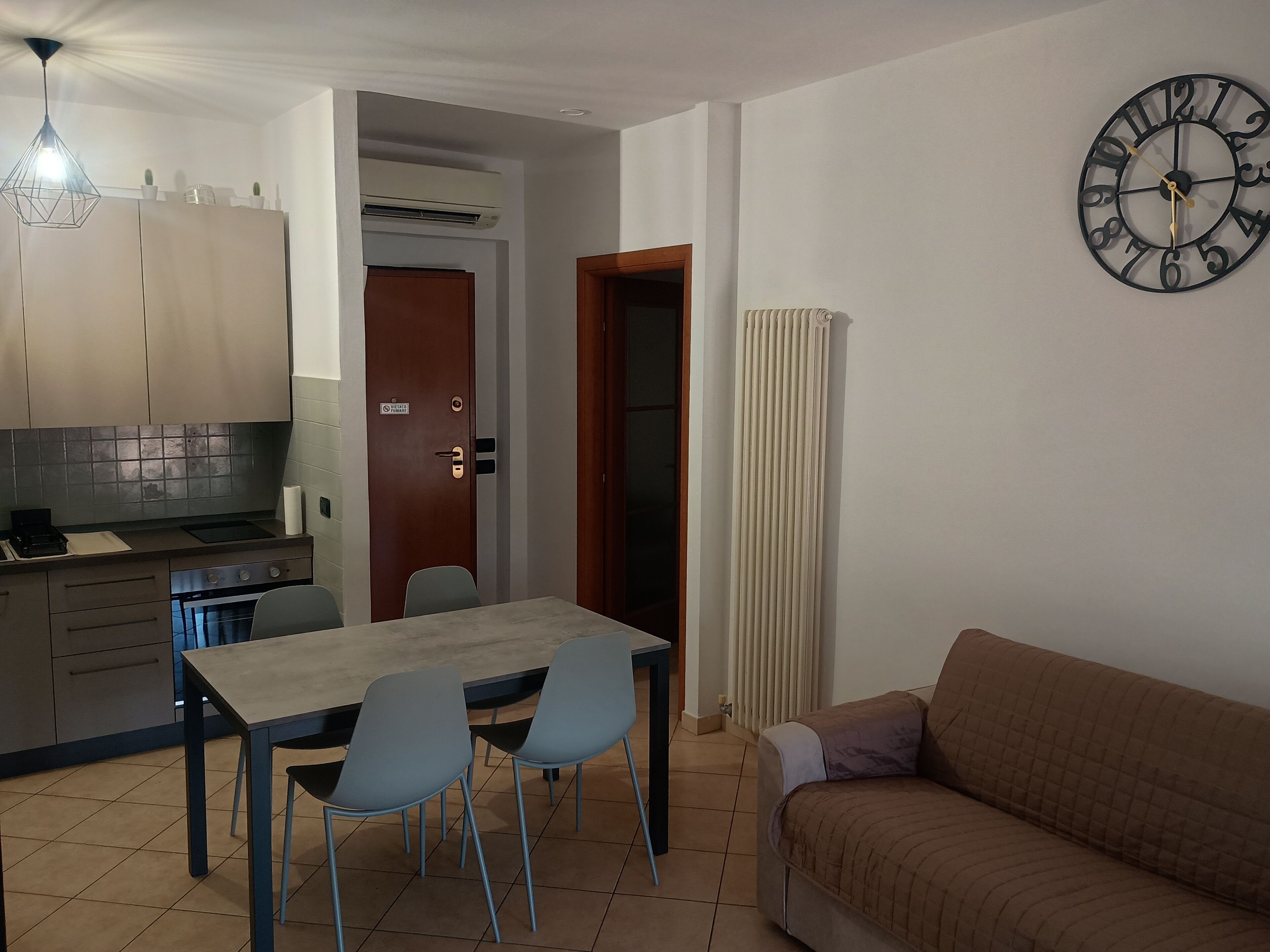A compact living space with a dining table, chairs, a sofa, a clock, and a kitchen area.