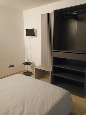 Een moderne slaapkamer met een bed, een aan de muur bevestigde televisie, een kledingkast en een tafeltje met een lamp.