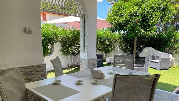 Un patio al aire libre con una mesa lista para dos personas, rodeado de sillas, una parrilla y vistas a una casa y follaje.