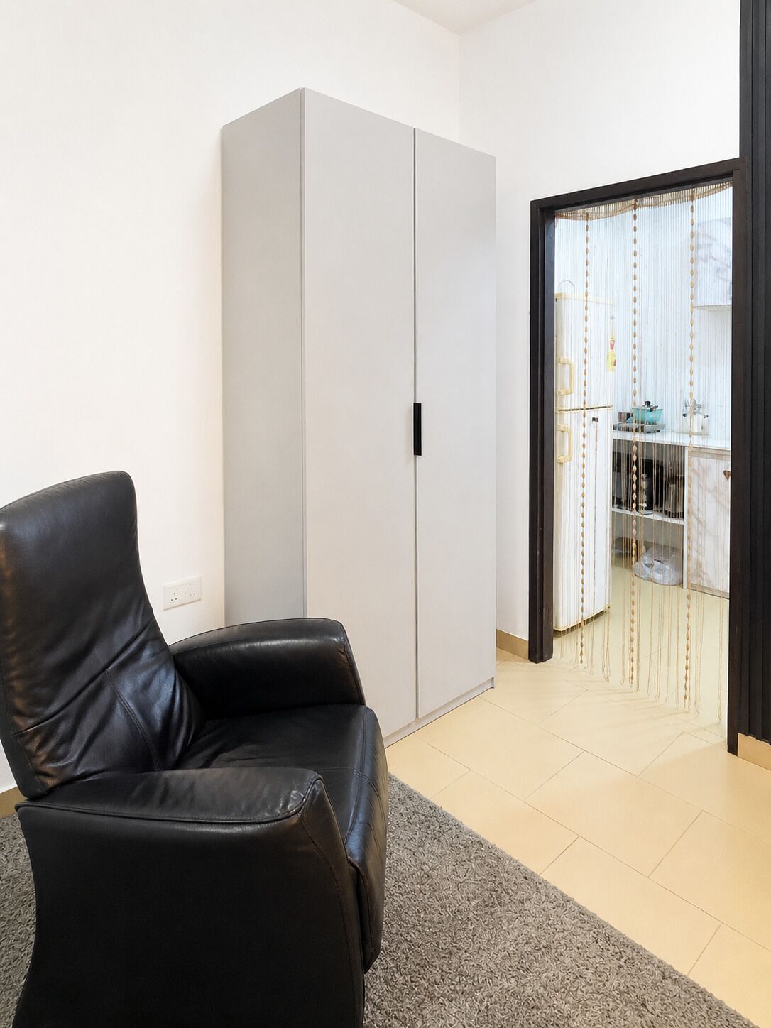 Una habitación con una silla de cuero negro, un armario blanco y una cocina visible a través de un vano.