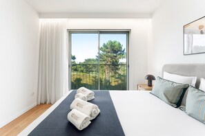 Habitació d'hotel amb un llit, una taula de nit, una làmpada, vista a la verdura a través d'una porta de balcó i una finestra amb vista als arbres.