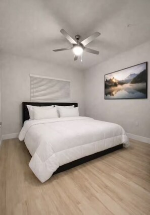 Un dormitorio con una cama, un ventilador de techo, una ventana con persianas y un cuadro en la pared.