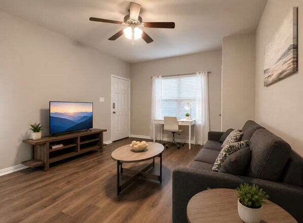 A living room with a television, a coffee table, a sofa, a ceiling fan, and a window with blinds.