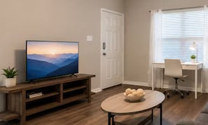 Ein Wohnzimmer mit einem Flachbildfernseher auf einem Holzboden, einem runden Tisch mit Obstschale, einem weißen Bürostuhl und einem Fenster mit Jalousien.
