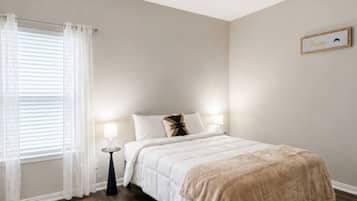 A bedroom with a bed, a bedside table, a lamp, a window with curtains, and a wall-mounted picture frame.