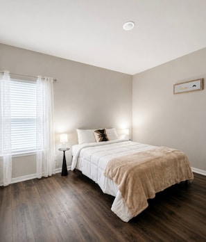 Ein Schlafzimmer mit einem Bett, einem Nachttisch, einer Lampe, einem Fenster mit Vorhängen und einem Bilderrahmen an der Wand.