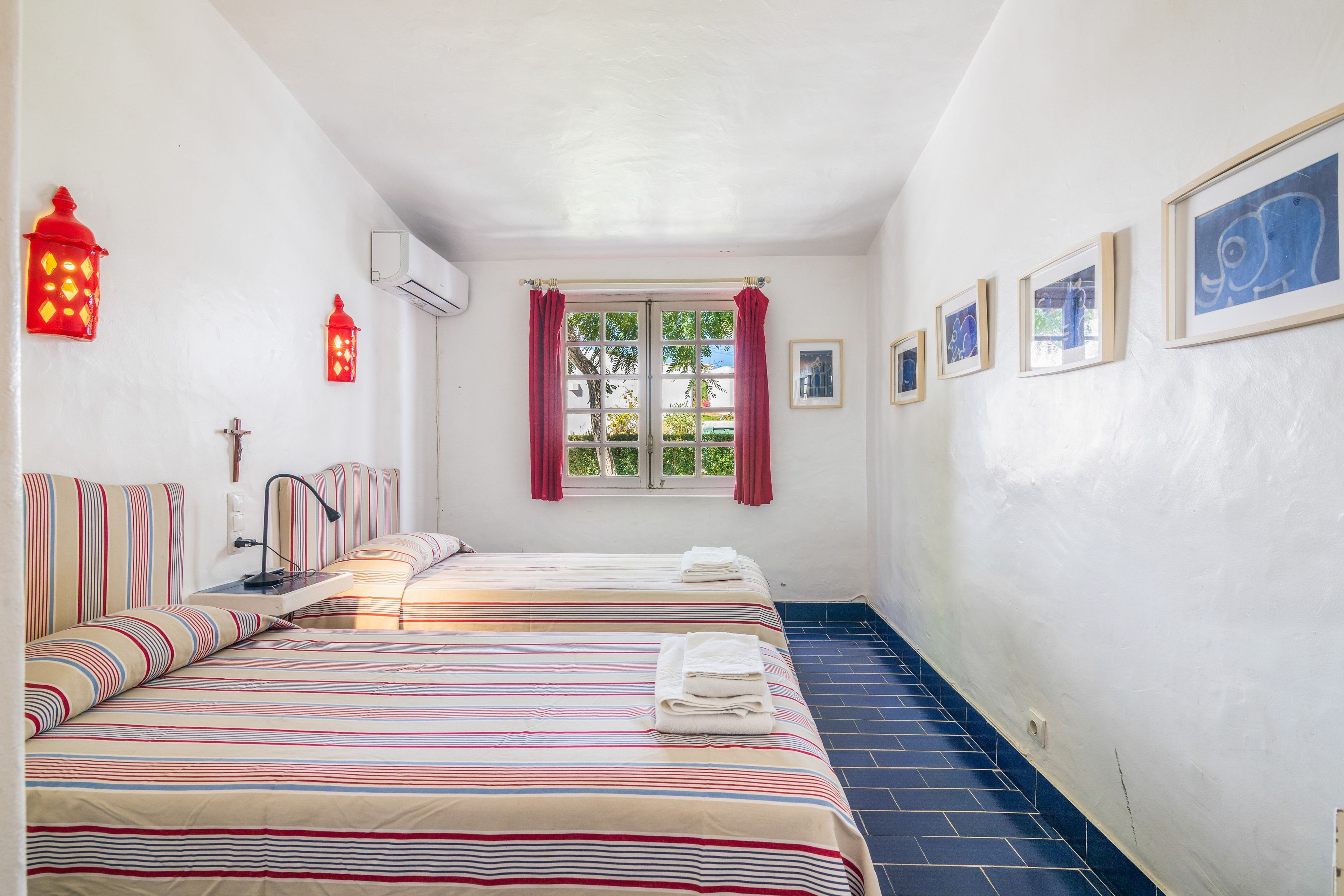 Une chambre d’hôtel avec deux lits, une fenêtre avec des rideaux, un climatiseur fixé au mur et des œuvres d’art encadrées.