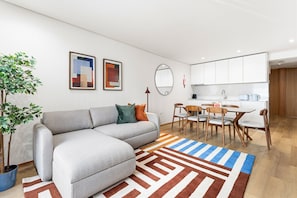 Ein modernes Wohnzimmer mit einem grauen Sofa, einem Essbereich mit Holzsesseln und einer Küche im Hintergrund.
