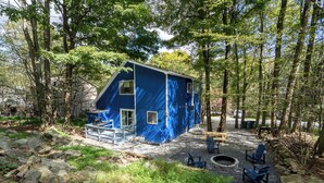 Una cabaña azul con terraza, rodeada de árboles, equipada con mobiliario exterior y un brasero.