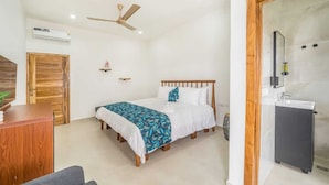 A bedroom with a bed, a desk, a wardrobe, a ceiling fan, an air conditioning unit, and a bathroom with a sink and mirror. - Crown Luxury Apartments  (Kokrobite)