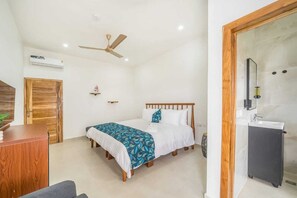 Une chambre comprenant un lit, un bureau, une armoire, un ventilateur de plafond, un appareil de climatisation et une salle de bain avec un lavabo et un miroir.