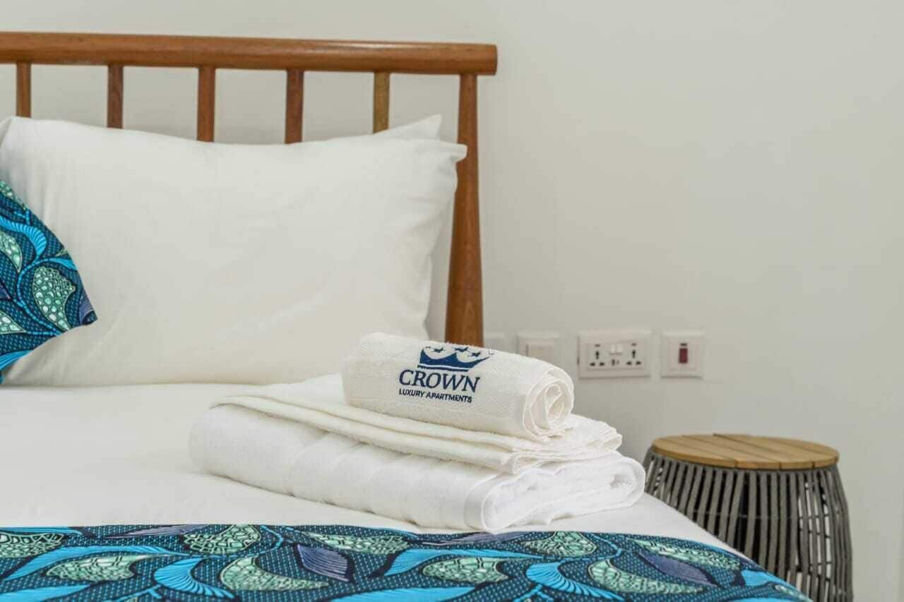 A neatly made bed with a white pillow, a rolled-up towel with 'CROWN LUXURY APARTMENTS' text, and a patterned bedspread.