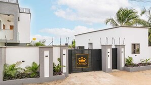 A gated entrance with a 'Crown' sign, a modern building facade, and a landscaped area with palm plants. - Crown Luxury Apartments  (Kokrobite)