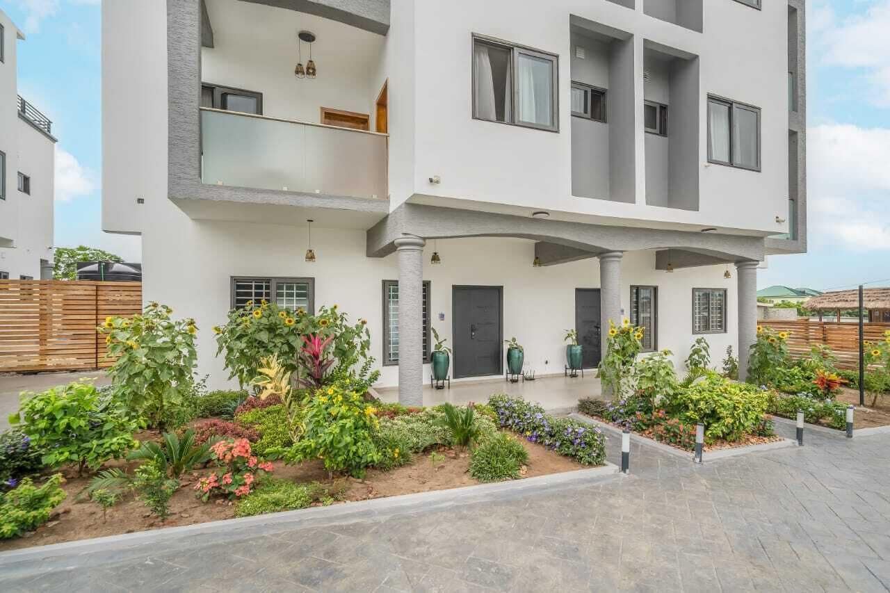 A modern two-story residential building with a balcony, a front garden with various plants, and a paved driveway.