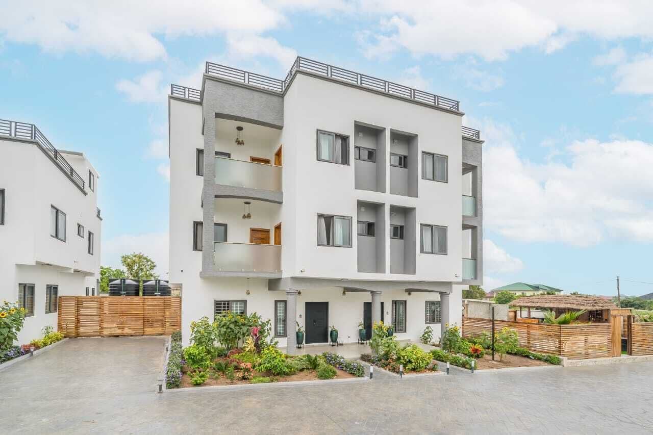 Un immeuble résidentiel moderne à plusieurs étages, doté de balcons, d’un espace extérieur couvert aménagé en coin salon et d’un jardin paysager.