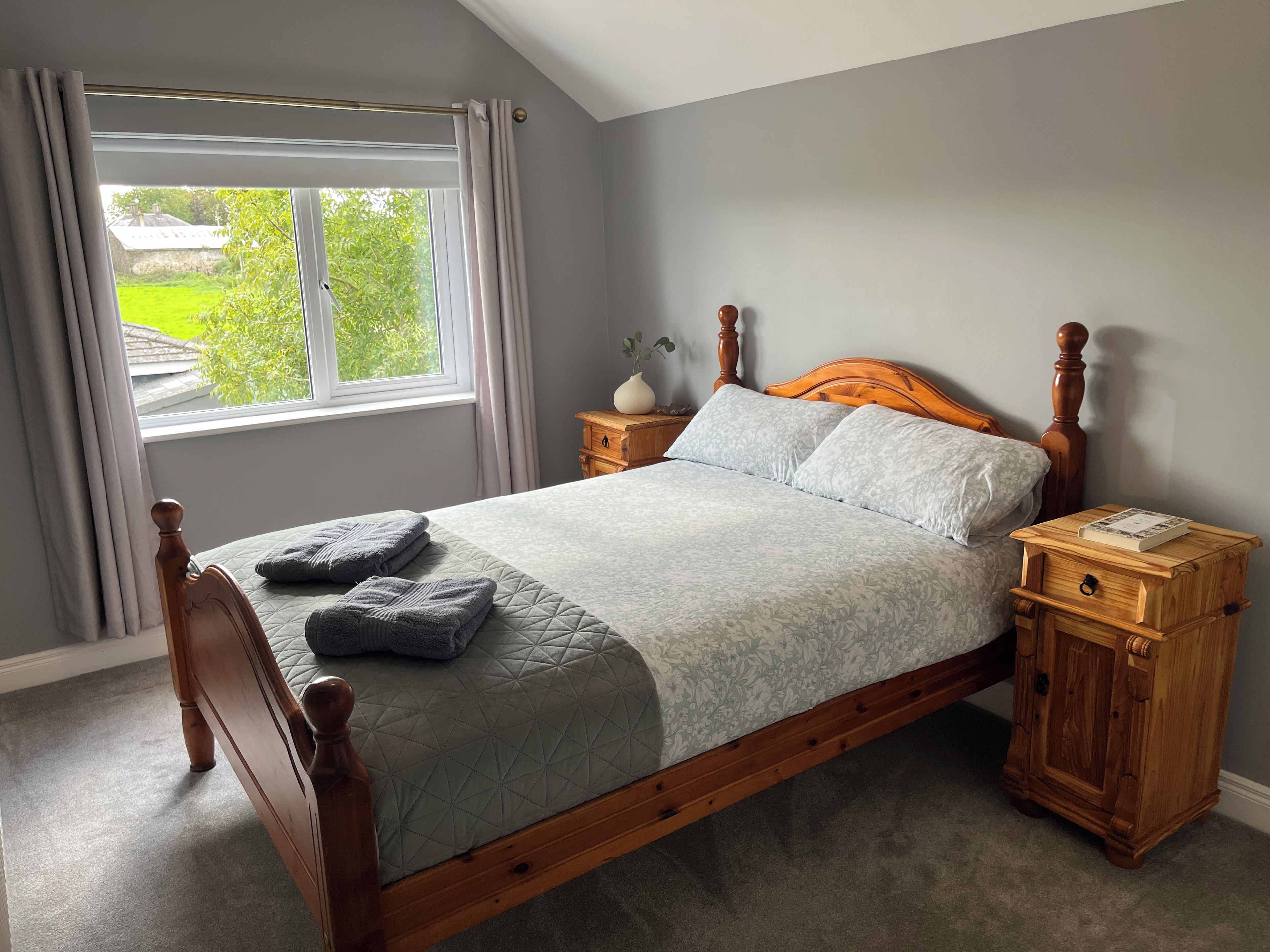 A neatly made bed with a wooden headboard and footboard, a bedside table with a book, and a window with curtains.