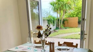 Een eetgedeelte met een tafel voor vier personen, stoelen, een vaas met bloemen en uitzicht op een tuin door een open deur.