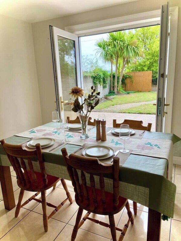A dining area with a table set for four, chairs, a vase with flowers, and a view of a garden through an open door.