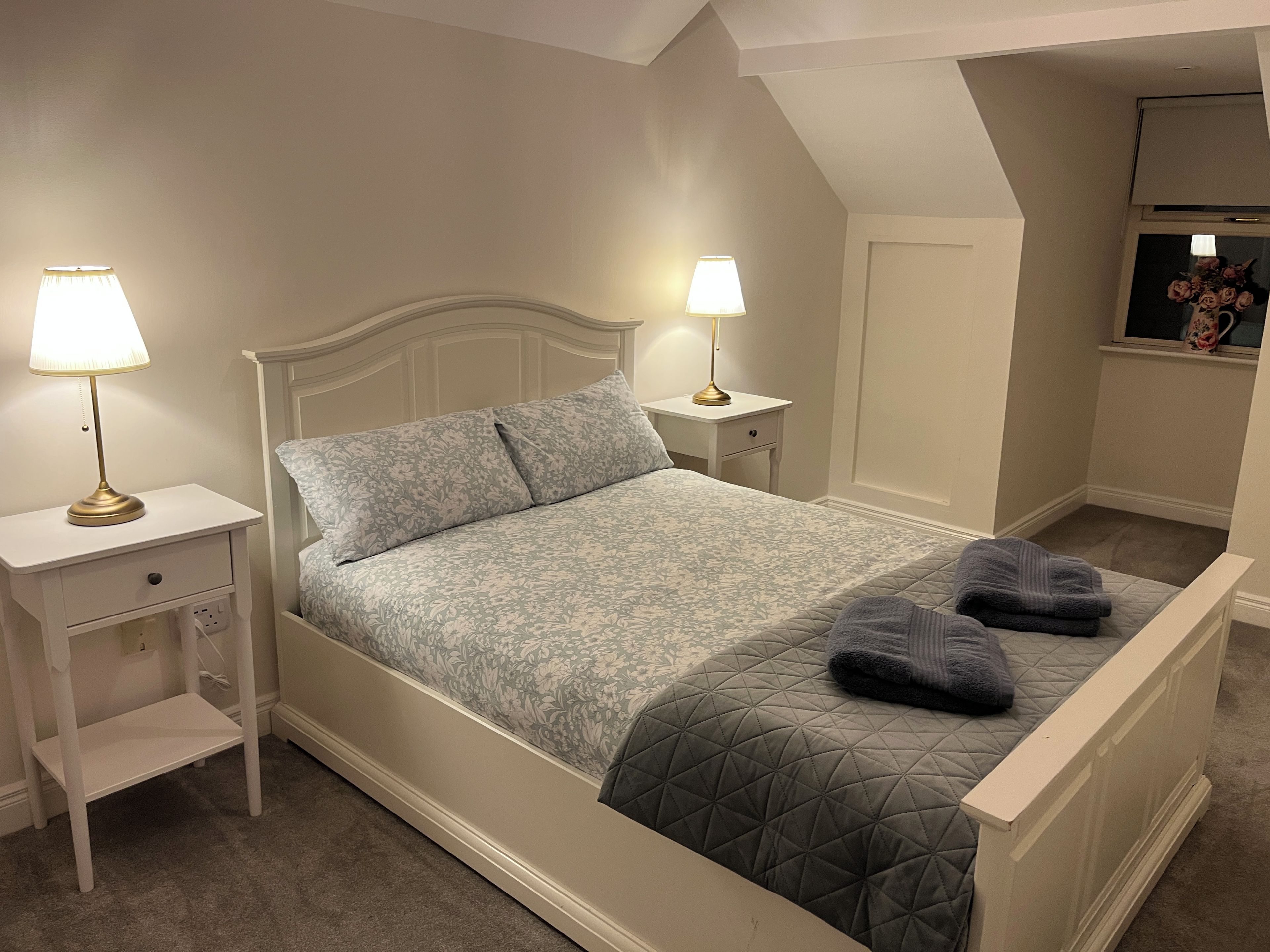 A bedroom with a bed, two bedside tables with lamps, a nightstand, and a window with a view.