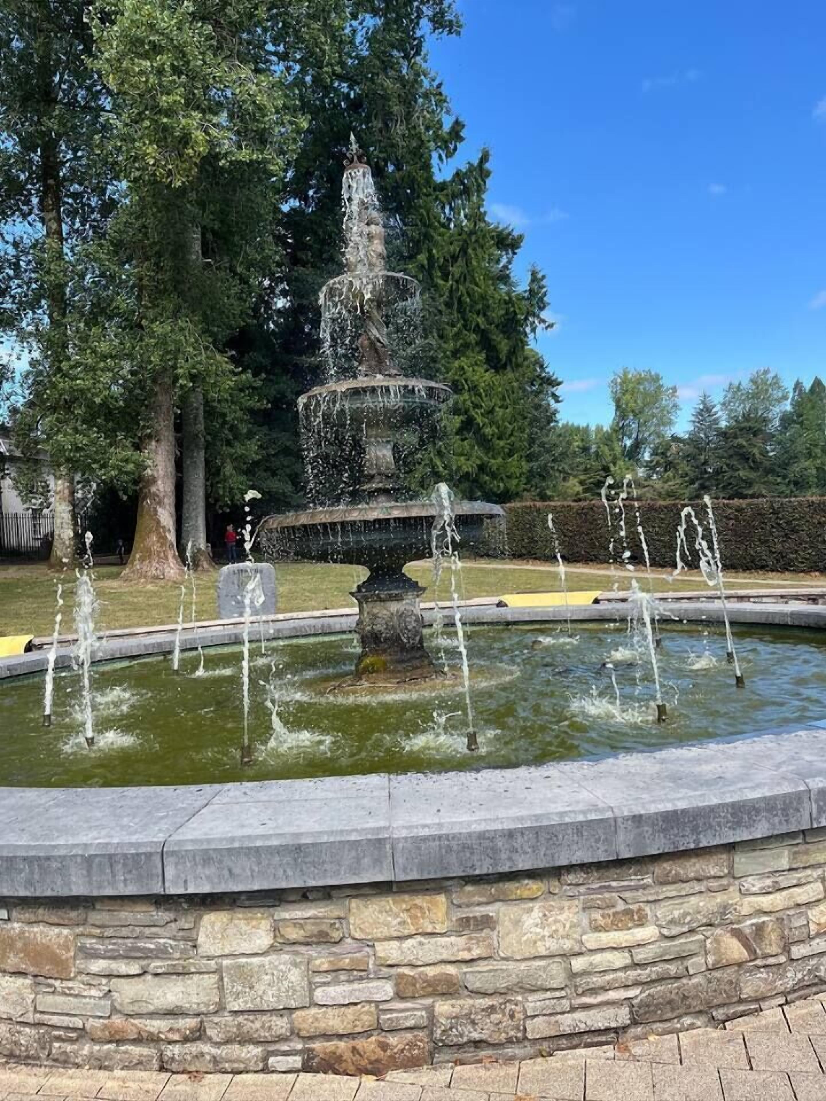 A multi-tiered fountain with water jets in a stone basin, surrounded by trees and a clear sky.