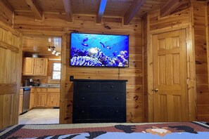 A wooden cabin interior with a television mounted above a black dresser, a kitchen area with wooden cabinets, and a patterned rug on the floor. - Mountain View Retreat 🏔️ Private Hot Tub, Arcade Room, Steps to Pigeon Forge Fun (Pigeon Forge)