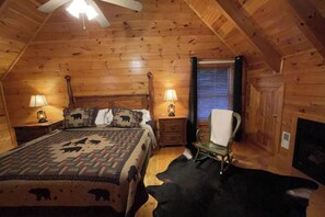 A log cabin bedroom with a bed, two bedside lamps, a chair, a window with blinds, and a fireplace. - Mountain View Retreat 🏔️ Private Hot Tub, Arcade Room, Steps to Pigeon Forge Fun (Pigeon Forge)