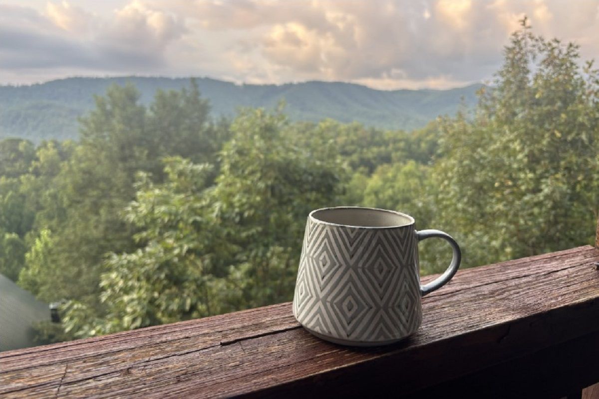 Una taza sobre un repisa de madera con vistas a una cadena montañosa boscosa.