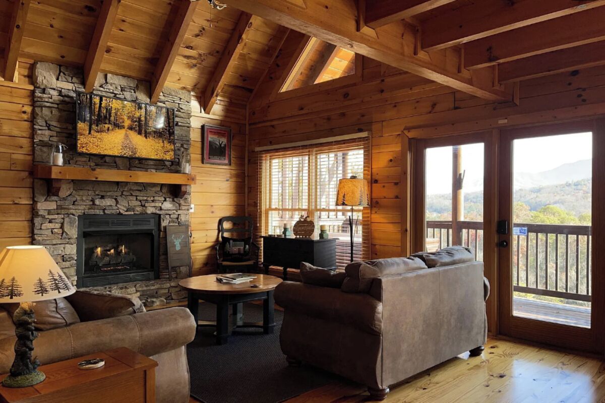 A cozy cabin interior with a stone fireplace, wooden beams, a sofa, a table, and a view through a door.