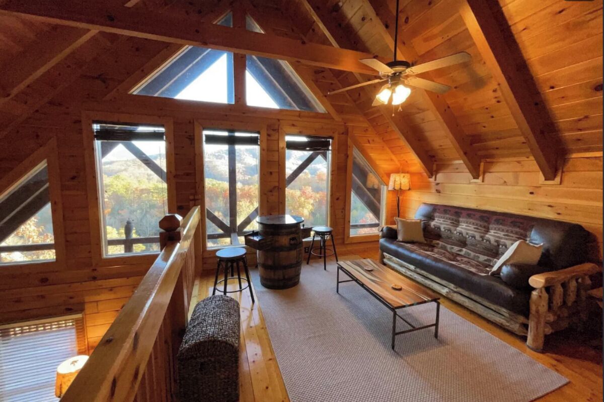 A cozy cabin with a loft-style ceiling, large windows, a wooden bench, a sofa, and a table.