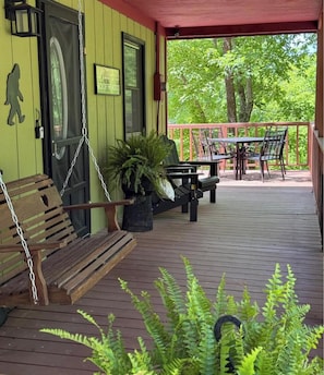 A wooden deck with a swing chair, a bench, and a table with chairs. There are potted plants and a sign on the wall.