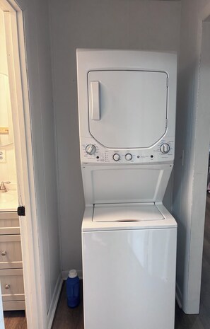 A compact, white, front-loading washing machine in a small laundry room with a door on the left and a blue detergent bottle on the floor.