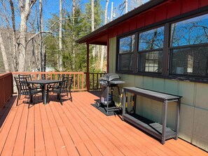 A wooden deck with a table and chairs, a grill, and a window overlooking a forest.