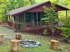 A cabin with a wooden deck, a covered porch, and a fire pit surrounded by rocks.
