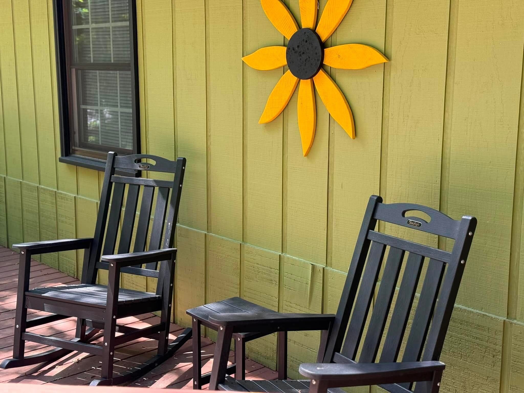 Twee houten stoelen met metalen poten, geplaatst op een houten terras tegen een gele muur met bloemendecoratie in geel en zwart.