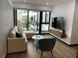 A modern living room with a sofa, a blue armchair, a coffee table, and a TV mounted on the wall. - 3-Bedroom Apartment with Heated Pool & Gym - Aparts4Days (San Isidro)