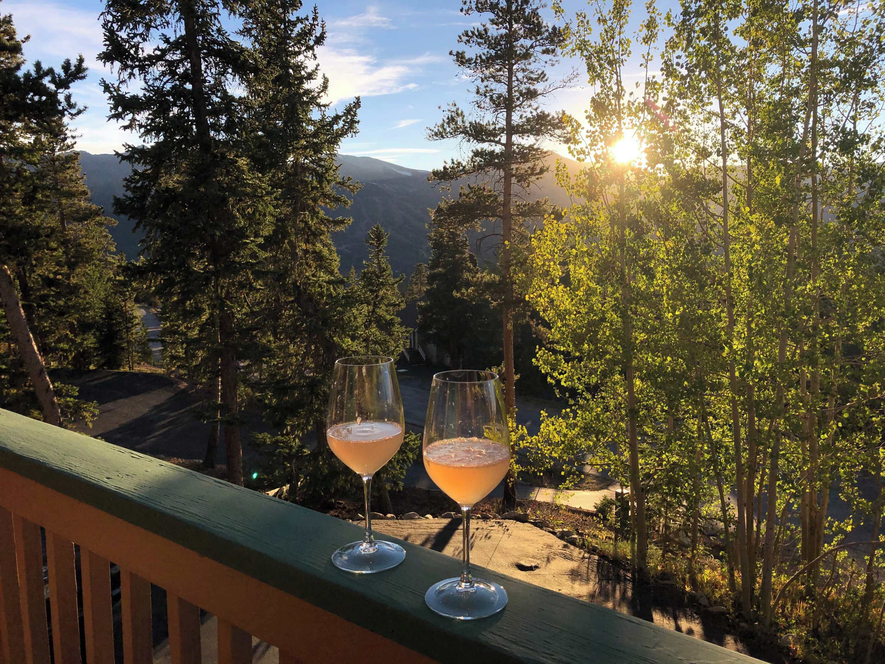 Two glasses of a beverage on a wooden deck with a view of a forest and mountains in the background.