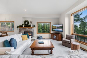 Ein modernes Wohnzimmer mit Kamin, großem Fernseher und Blick auf einen Wald durch die Fenster.