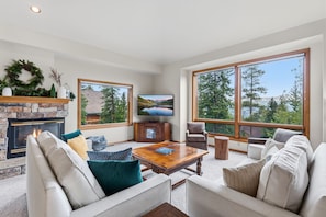A spacious living room with a fireplace, comfortable seating, a coffee table, and large windows offering scenic views. - First Light Cabin Breck - Deck & Views (Breckenridge)