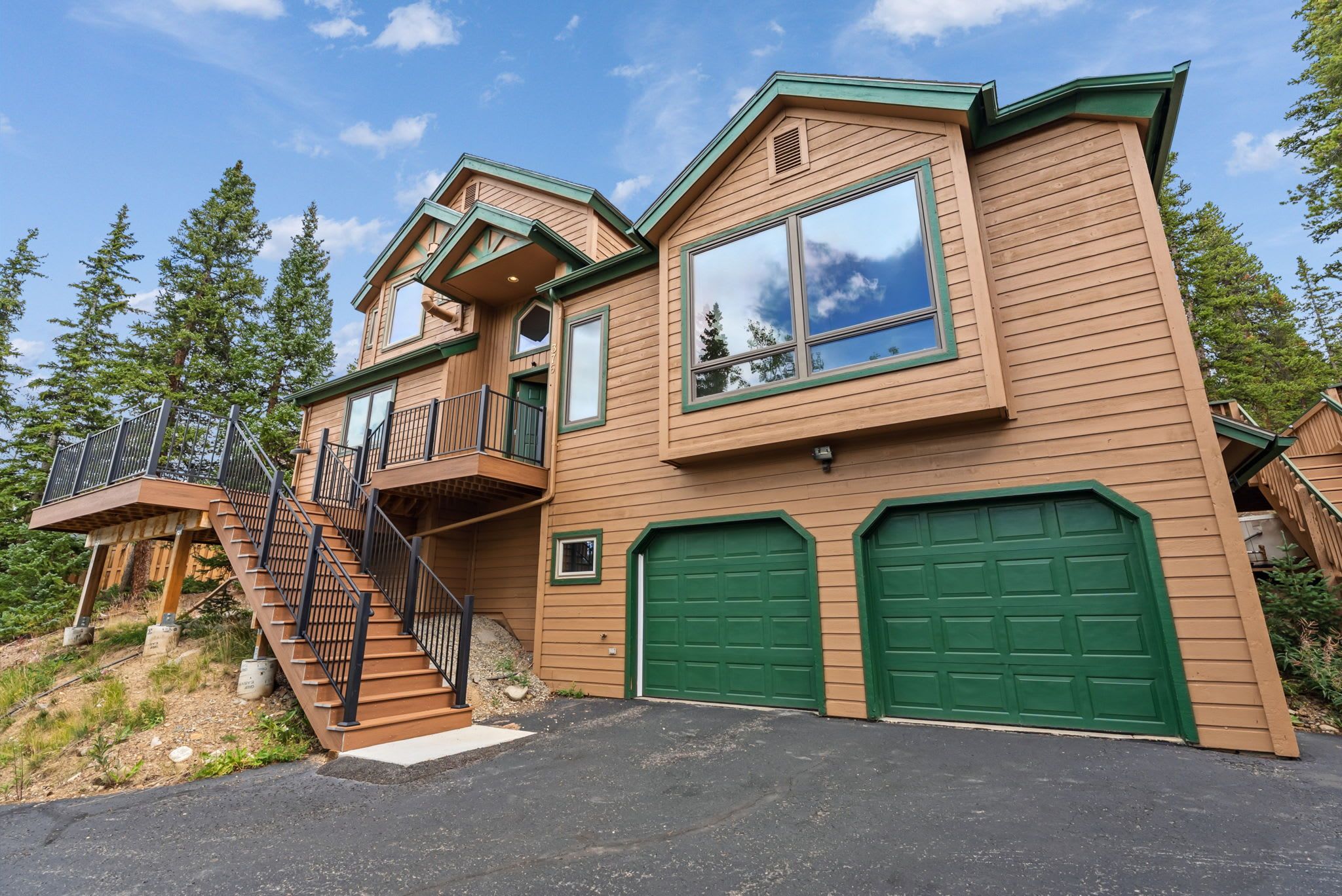A two-story wooden house with a balcony, a staircase, and two green garage doors.