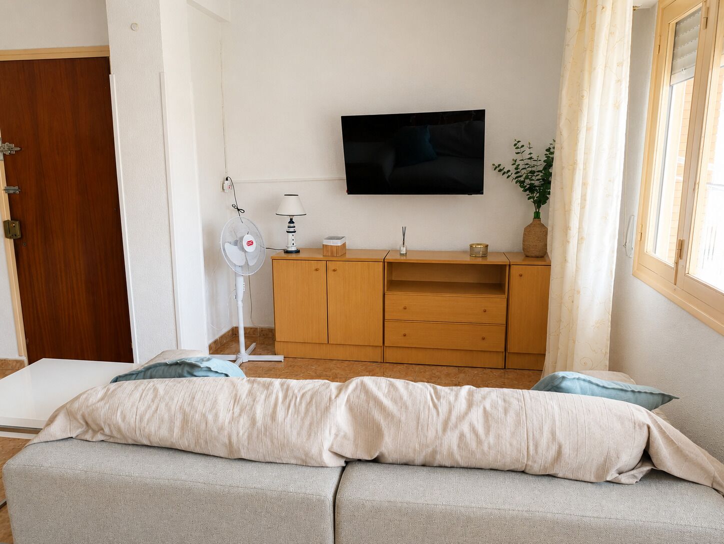 Una sala de estar con un sofá, un televisor montado en la pared, un mueble de madera, un ventilador de pie y una ventana con cortinas.