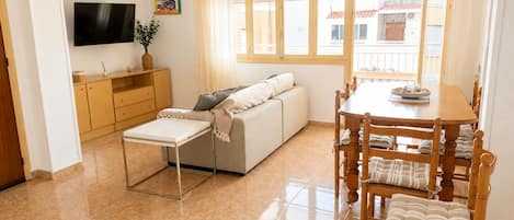 Una sala de estar con un sofá, una mesa de comedor con sillas, un televisor montado en la pared y un ventanal con cortinas.