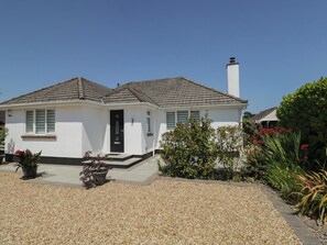 Eenlaags huis met een witte gevel, een zwarte voordeur en een schoorsteen. Er is een grindpad en een kleine tuin met verschillende planten en bloemen.