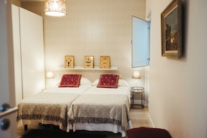 A hotel room with two beds, a small table, a wall-mounted shelf with framed pictures, and a door. - Vrbo Property (Sevilla)