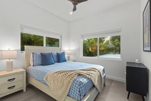 Ein Schlafzimmer mit einem Bett, Nachttischen, einer Lampe, einem Deckenventilator und Fenstern mit Blick auf Grünflächen.
