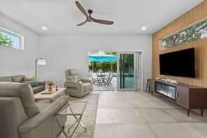 A modern living room with a sofa, armchair, TV, fireplace, ceiling fan, and a view through a glass door to an outdoor patio. - Modern 3BR • Private Backyard • Minutes to Downtown & Siesta Key (Sarasota)